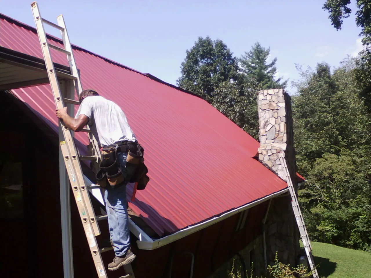 Roofer on a ladder beside a red metal roof and stone chimney — Soffit & Fascia Repair by Roofing Brothers in Winchester